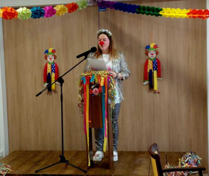 A person in a shiny outfit stands at a microphone, reading from a paper, with clown decorations and colorful streamers in the background.
